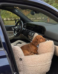 A brown dachshund dog rests inside a cream-colored, sherpa fleece car seat positioned in the open passenger door of a dark blue car.