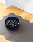 Blue ceramic bowl with pet food on a gray mat with 'Le Noor' branding.