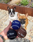Three dog toy balls held in a hand in front of two French bulldogs in a garden setting. The balls are textured and printed with the brand name 'Le Noof'.