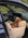 A brown dachshund dog rests inside a cream-colored, sherpa fleece car seat positioned in the open passenger door of a dark blue car.